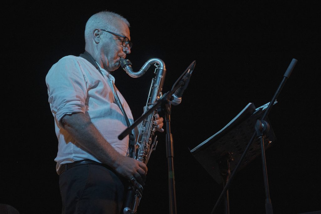 Musicista sul palco durante un concerto serale, mentre suona un sassofono davanti a microfoni e leggio. La scena è illuminata da luci fredde su sfondo scuro, con atmosfera raccolta e intensa.
