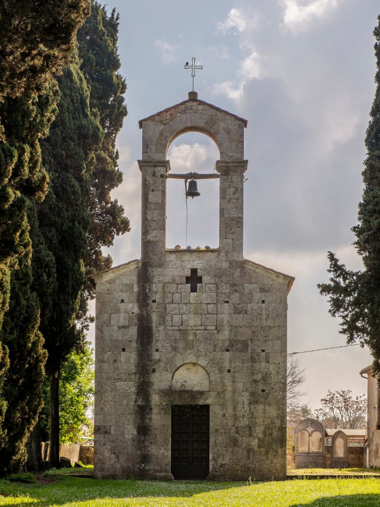Facciata in pietra della chiesa di San Miniato in Marcianella a Cascina, con campanile a vela, campana, croce sommitale e cipressi ai lati.
